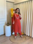 Woman wearing a rust red straight kurta set with white floral embroidery, festive indoor setting with marigold flowers.