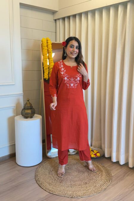 Woman wearing a rust red straight kurta set with white floral embroidery, festive indoor setting with marigold flowers.