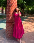Woman wearing a magenta flared designer kurti set with intricate neckline embroidery, paired with matching pants, standing outdoors against a brick pillar – ideal for festive and party wear.