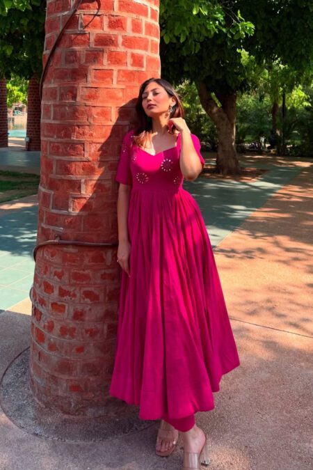 Woman wearing a magenta flared designer kurti set with intricate neckline embroidery, paired with matching pants, standing outdoors against a brick pillar – ideal for festive and party wear.