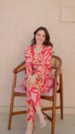 Woman wearing a vibrant red and pink designer printed kurti set with abstract floral patterns, gold lace detailing on neckline and cuffs, paired with matching straight pants