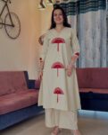 Woman wearing a beige cotton kurta pant set with red fan-style embroidery, standing indoors in a cozy ethnic look perfect for casual or daily wear.