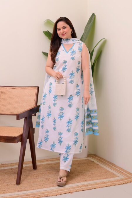 Woman wearing a white and blue floral printed sleeveless kurta with matching pants and a dupatta, standing in a modern indoor setup with a wooden chair.