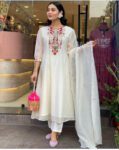 Woman wearing a white embroidered kurta set with dupatta and pants, holding a pink potli bag and posing stylishly outside a designer boutique.
