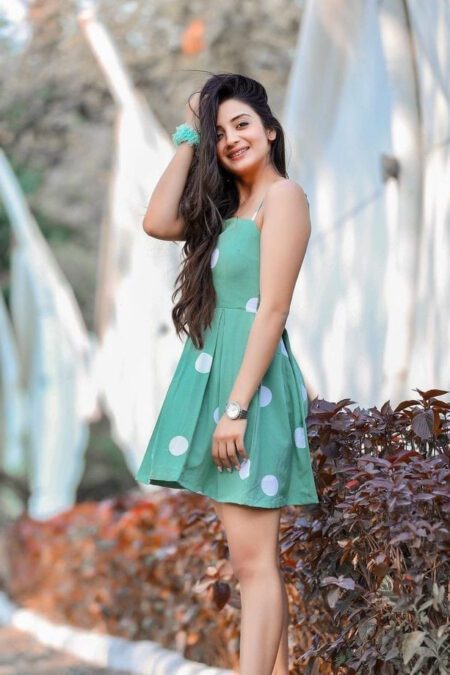 Young woman wearing a green polka dot strappy short dress, standing outdoors, smiling confidently in a stylish casual look.