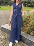 Woman wearing a navy blue sleeveless cotton kurti-style top with floral embroidery, paired with wide-leg palazzo pants, holding a woven handbag in a garden setting.