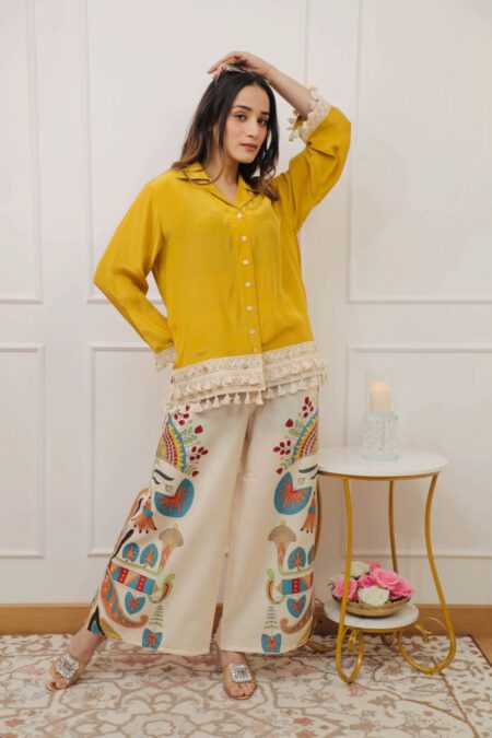 Woman wearing a mustard button-down shirt with lace trim and colorful ethnic-printed palazzo pants in a co-ord set, posing indoors with minimal decor.