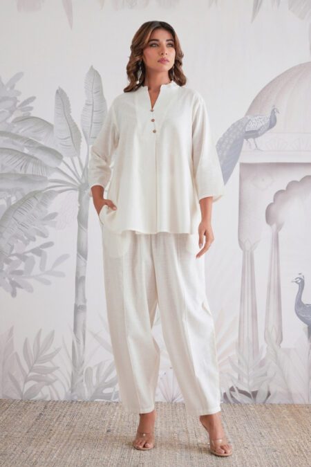 Woman wearing a white full-sleeve co-ord set for women with matching cotton tapered pants, standing confidently against a traditional artistic backdrop.